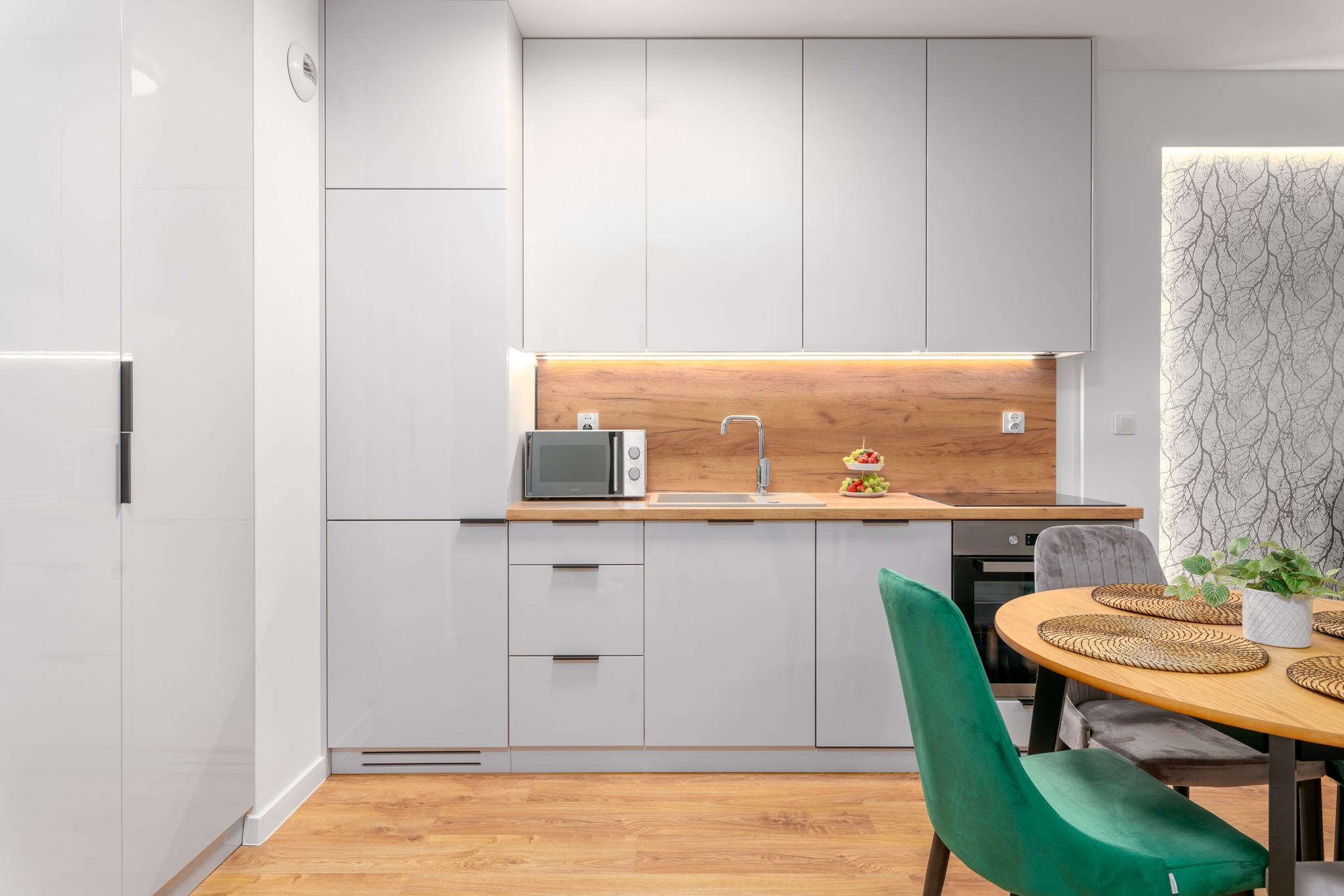 Nowoczesna, jasna kuchnia z białymi, błyszczącymi szafkami, drewnianym blatem i okrągłym stołem z zielonym krzesłem.