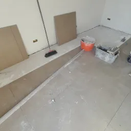 Przygotowania do ułożenia płytek podłogowych: widoczne płyty MDF, poziomica, wiadra z zaprawą, szpachelka, krzyżyki dystansowe i szczotka.