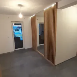 Jasne wnętrze domu w trakcie wykończenia, z szarymi płytkami na podłodze, drewnianą ścianką działową z pionowych listew z oświetleniem LED i widokiem na ogród przez drzwi balkonowe.