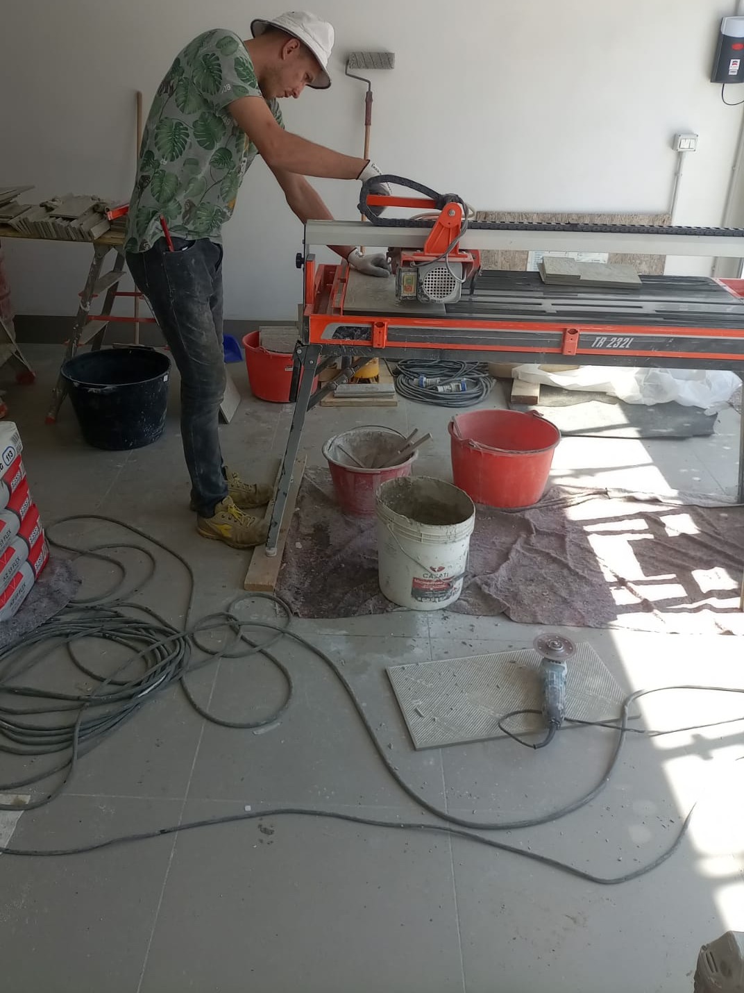 Pracownik w kapeluszu tnie płytki ceramiczne na mokro przy użyciu przecinarki elektrycznej, widoczne narzędzia, wiaderka, worki z klejem i szlifierka kątowa na podłodze.