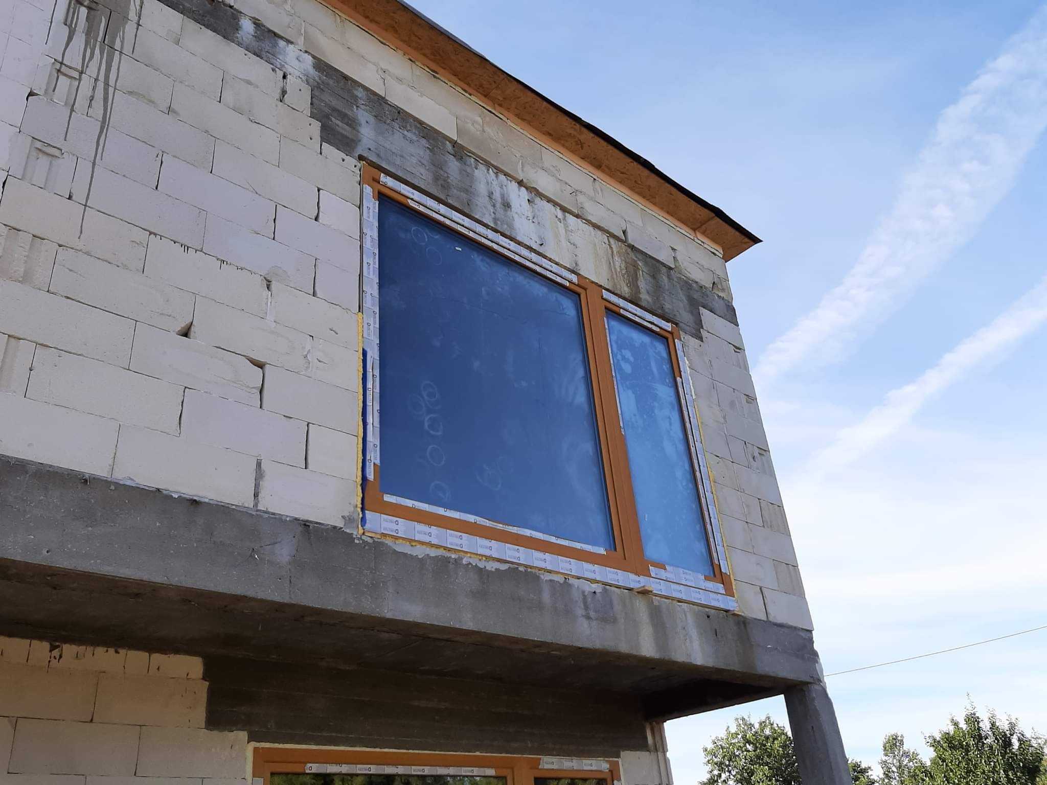 Surowy stan budynku z białych bloczków z zamontowanymi oknami z brązowymi ramami, zabezpieczonymi folią ochronną, widok na błękitne niebo z chmurami.