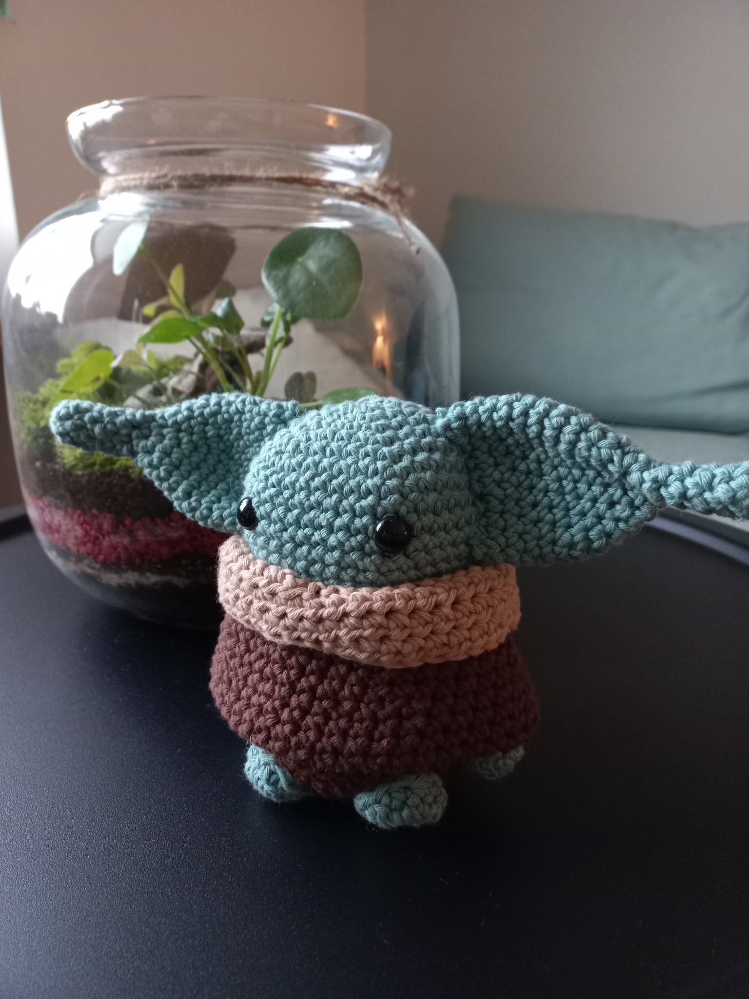 Amigurumi Baby Yoda w kolorach turkusowym, brązowym i beżowym, ustawiony obok szklanego słoja z roślinami, na czarnym stole.