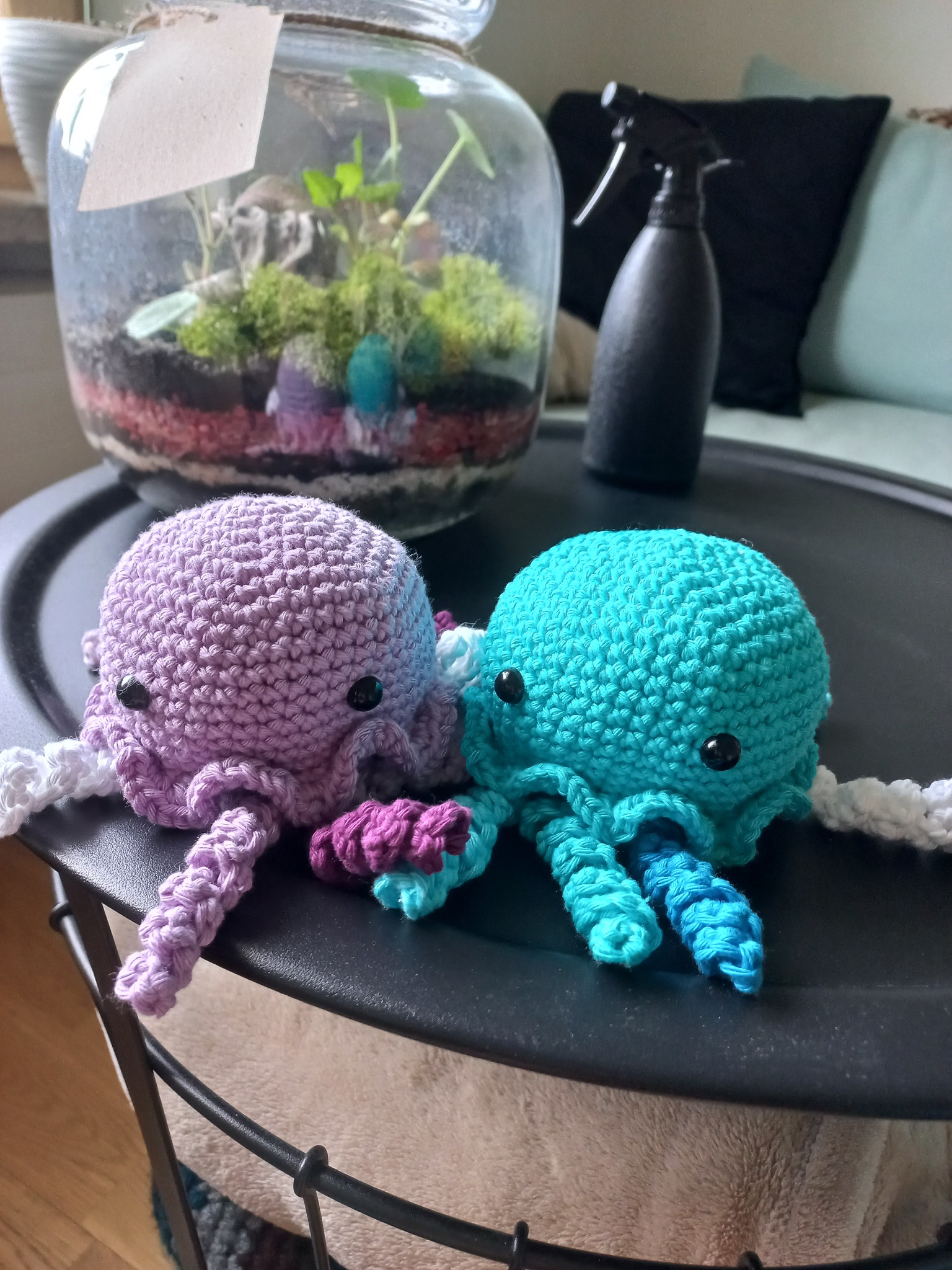 Dwie ręcznie robione na szydełku maskotki w kształcie meduz: fioletowa z różowymi mackami i turkusowa z niebieskimi, leżą na czarnym stoliku obok szklanego terrarium i czarnego spryskiwacza.