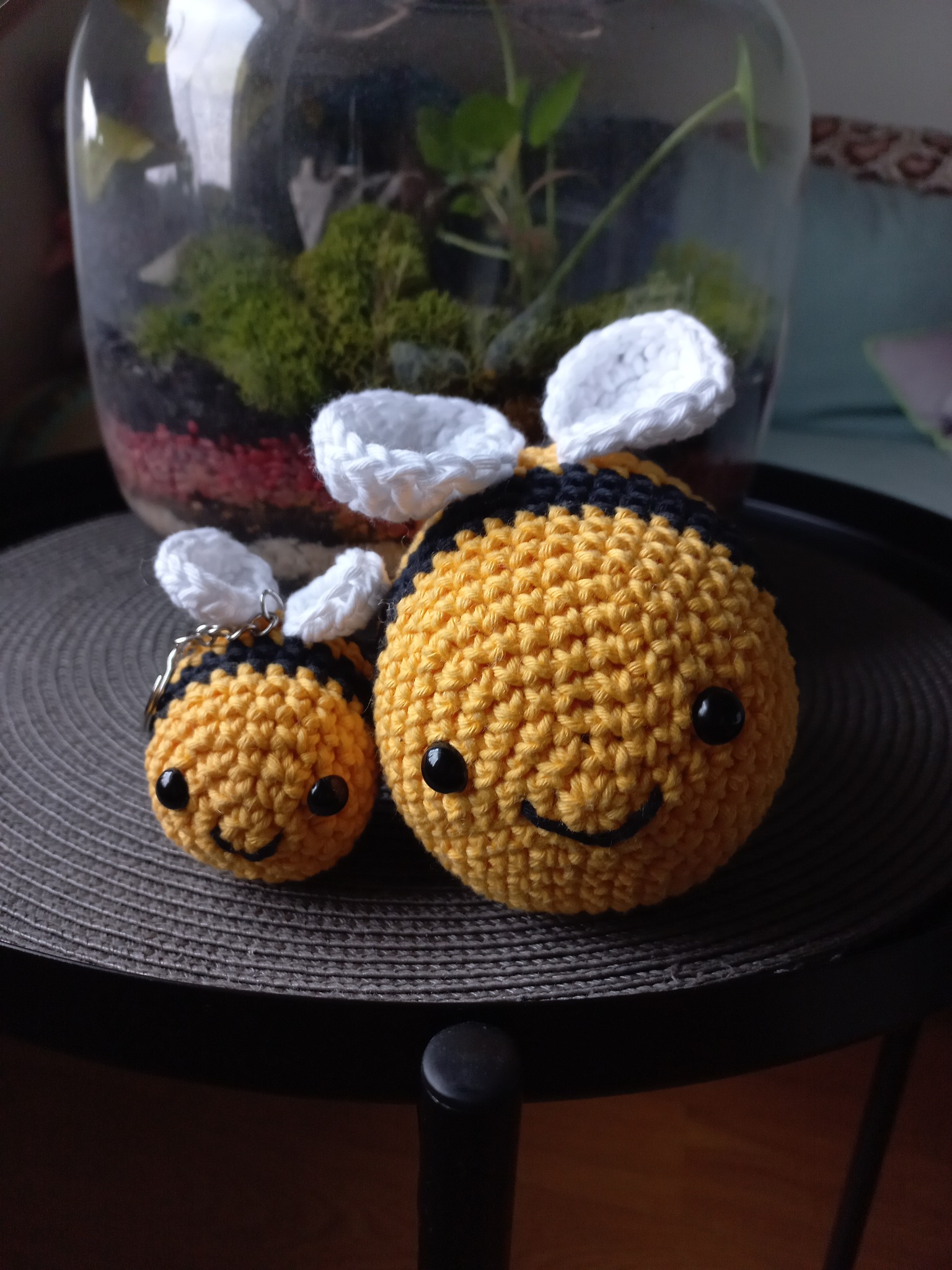 Dwie szydełkowane pszczółki: większa i mniejsza breloczek, ustawione na szarym, okrągłym podkładce na stole, w tle terrarium z roślinami i czerwonym piaskiem.