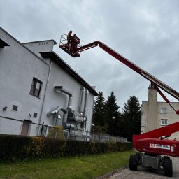 Ur serwis sp. Z o.o. - Pracownicy na podnośniku koszowym montują system wentylacyjny na elewacji budynku. Widoczne rury wentylacyjne i fragment elewacji. Pochmurne niebo w tle.