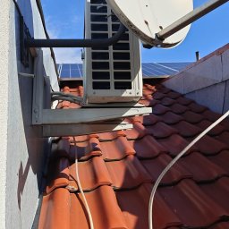 Ur serwis sp. Z o.o. - Biała jednostka zewnętrzna klimatyzacji zamontowana na dachu z czerwonej dachówki, obok antena satelitarna i panele słoneczne w tle, błękitne niebo.