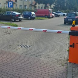 Ur serwis sp. Z o.o. - Automatyczna bariera parkingowa CAME z pomarańczową obudową i biało-czerwonym ramieniem, na tle parkingu z zaparkowanymi autami i budynkiem mieszkalnym.