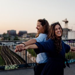 Magda Maroni Fotografia - Uśmiechnięta para, mężczyzna i kobieta, stoją plecami do siebie na moście z rozpostartymi ramionami, w tle miejski krajobraz i zachodzące słońce.