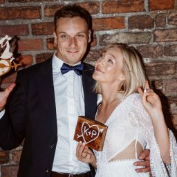 Magda Maroni Fotografia - Uśmiechnięta para młoda pozuje na tle ceglanej ściany, trzymając drewniany topper z sylwetką pary i globusem oraz drewniane serce z inicjałami K+P.