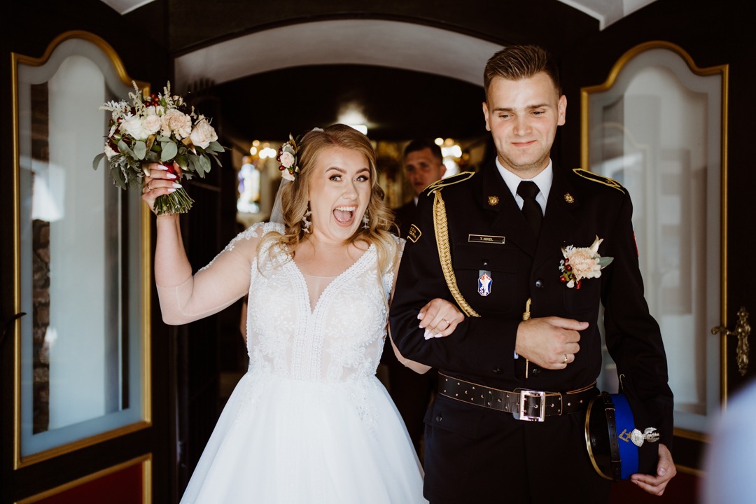 Uśmiechnięta panna młoda w koronkowej sukni, z bukietem w ręku, wychodzi z kościoła pod rękę z ubranym w galowy mundur strażaka panem młodym.