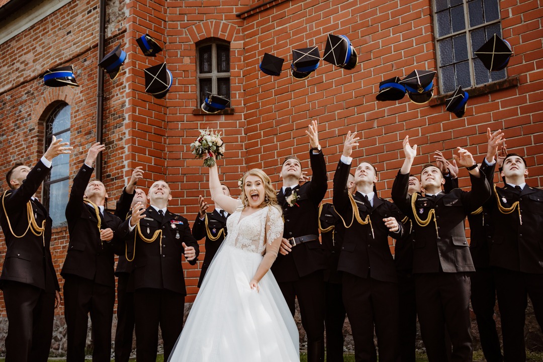 Uśmiechnięta panna młoda w białej sukni z bukietem i grupa mężczyzn w galowych mundurach strażackich rzucających czapki w górę na tle ceglanego budynku.