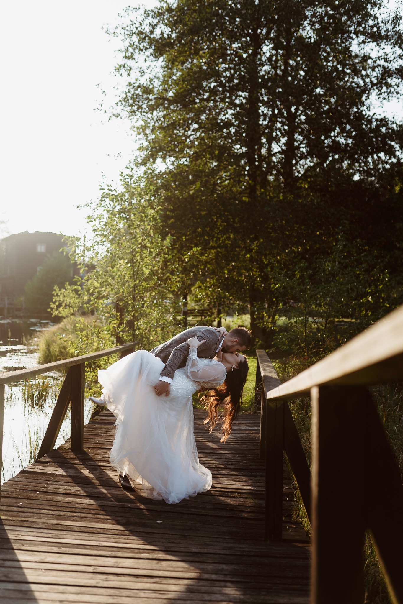 Panna młoda w białej sukni ślubnej, unoszona przez pana młodego w szarym garniturze podczas pocałunku na drewnianym moście w otoczeniu drzew i jeziora.
