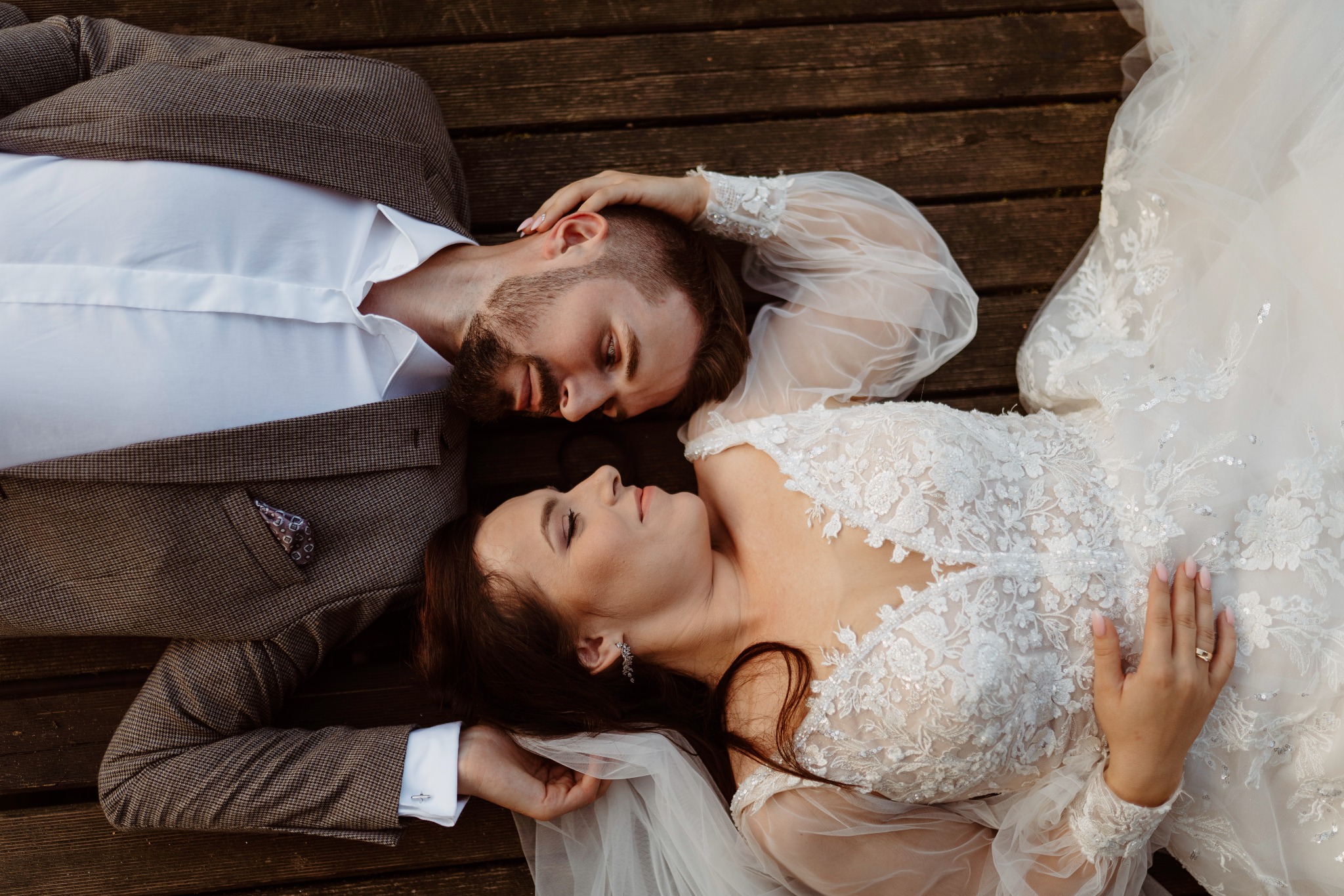 Młoda para leżąca na drewnianym pomoście, panna młoda w koronkowej sukni z długim rękawem i welonem, pan młody w brązowym tweedowym garniturze z poszetką w kieszonce, zbliżenie na twarze.