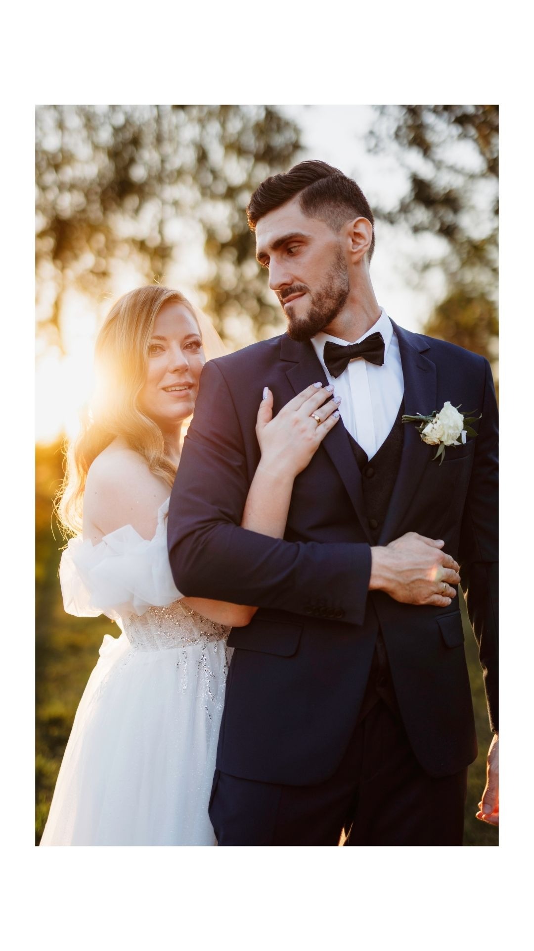 Para młoda: panna młoda w białej sukni z odkrytymi ramionami i pan młody w granatowym garniturze z muchą, obejmujący się na tle zachodzącego słońca.