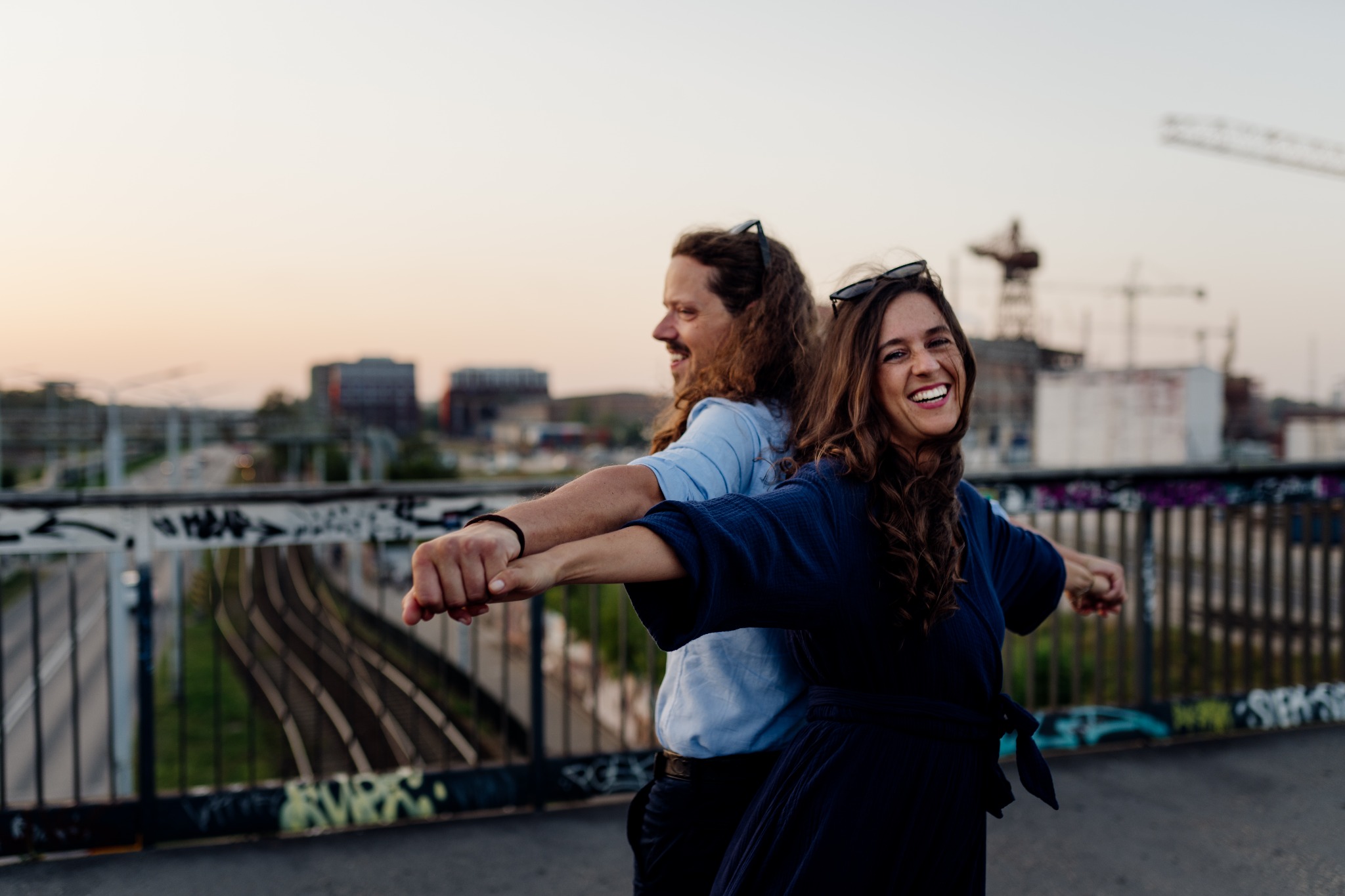 Uśmiechnięta para, mężczyzna i kobieta, stoją plecami do siebie na moście z rozpostartymi ramionami, w tle miejski krajobraz i zachodzące słońce.
