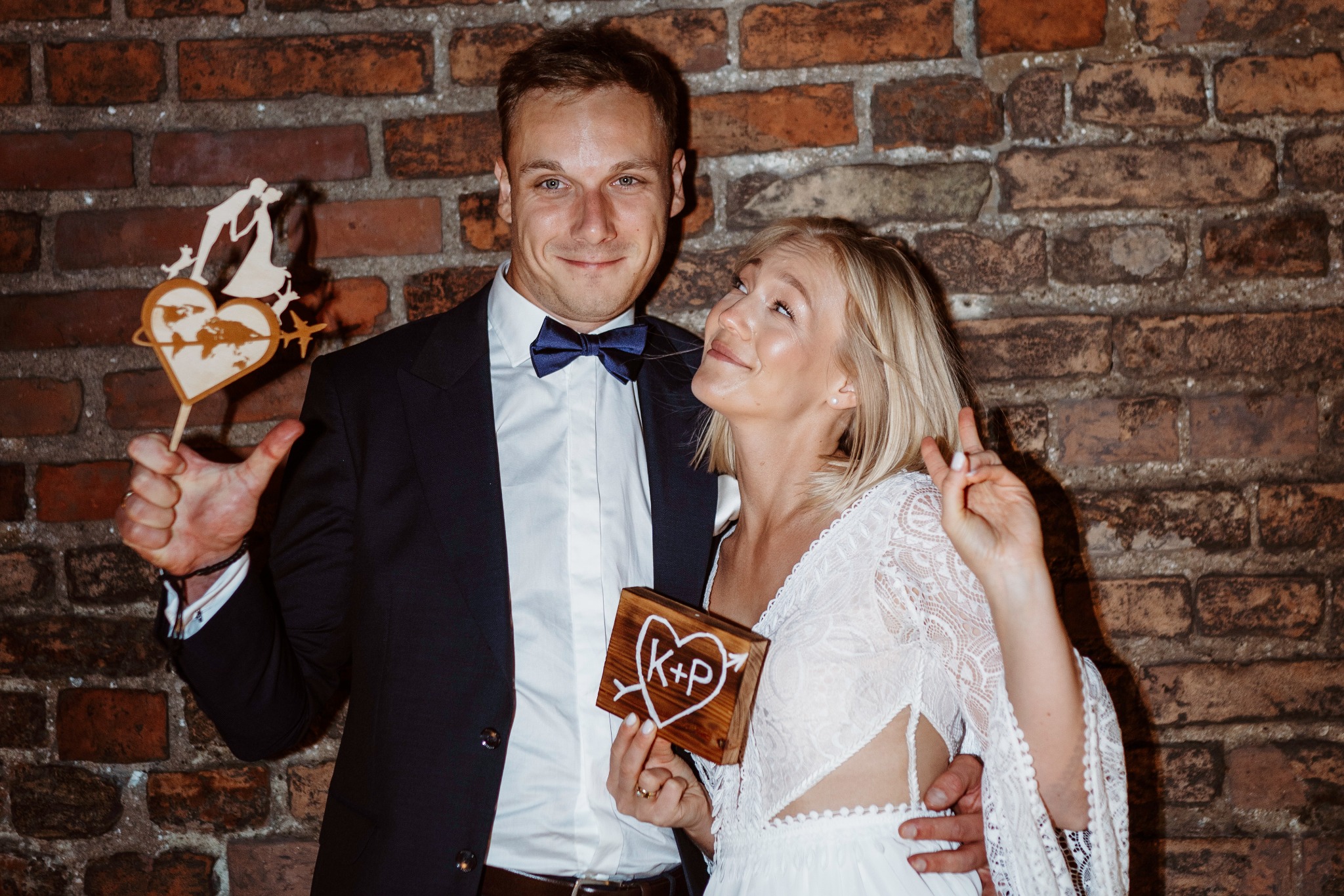 Uśmiechnięta para młoda pozuje na tle ceglanej ściany, trzymając drewniany topper z sylwetką pary i globusem oraz drewniane serce z inicjałami K+P.