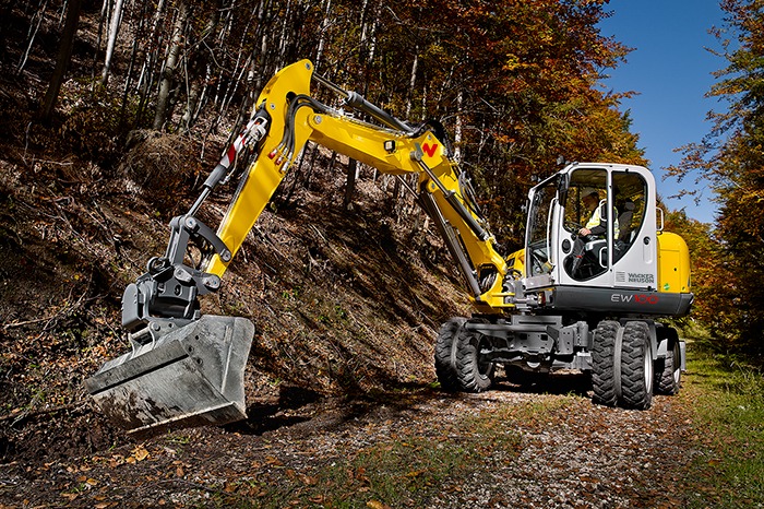 Żółta koparka kołowa Wacker Neuson EW100 na leśnej ścieżce, operator w kabinie, jesienne drzewa w tle.