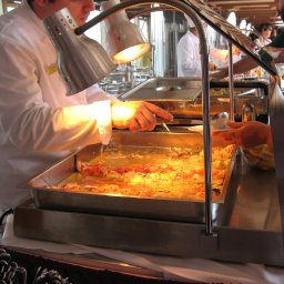 MEGAR Sp. z o.o. - Szef kuchni w białym uniformie serwuje danie z podgrzewacza bufetowego, oświetlonego lampami, z widocznymi kawałkami pomidorów i żółtym sosem, podczas obsługi gości.