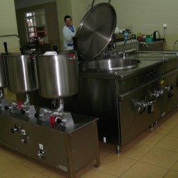 MEGAR Sp. z o.o. - Profesjonalna kuchnia ze stali nierdzewnej: kotły warzelne i stół warzelny z otwartą pokrywą, widoczna osoba w fartuchu w tle.