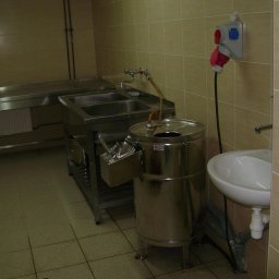 MEGAR Sp. z o.o. - Wnętrze zaplecza kuchennego z metalowym stołem, dwukomorowym zlewem, okrągłą maszyną ze stali nierdzewnej z kranem i białą umywalką, przyłącze elektryczne na ścianie, wyłożone beżowymi płytkami.