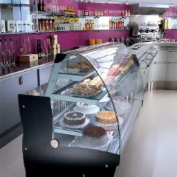 Nowoczesna, przeszklona witryna chłodnicza wypełniona ciastami i deserami w cukierni lub kawiarni; w tle widoczne półki z alkoholami i ekspres do kawy.