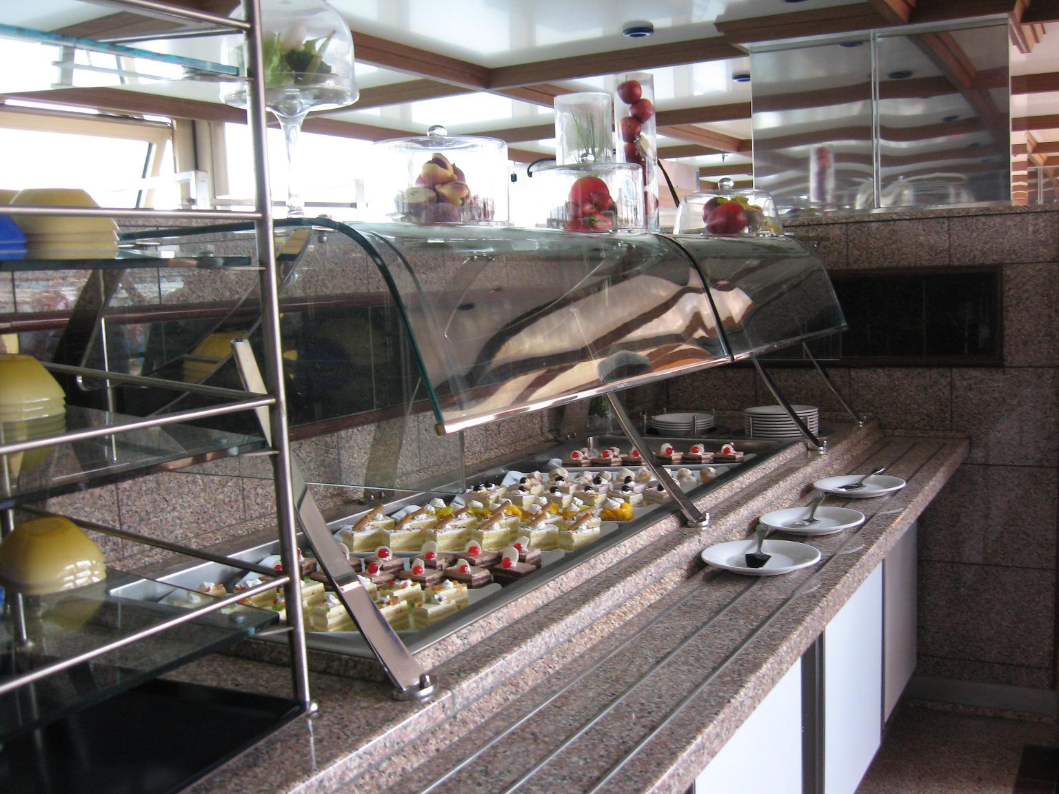 Szklana witryna chłodnicza z ekspozycją różnorodnych ciast i deserów na metalowych tacach, z talerzami i sztućcami w pobliżu, ustawiona w pomieszczeniu z wykończeniem z kamienia i lustrzanym sufitem.