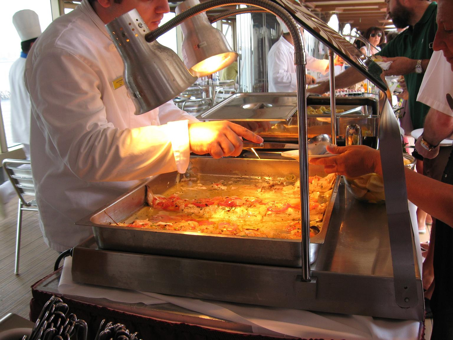 Szef kuchni w białym uniformie serwuje danie z podgrzewacza bufetowego, oświetlonego lampami, z widocznymi kawałkami pomidorów i żółtym sosem, podczas obsługi gości.