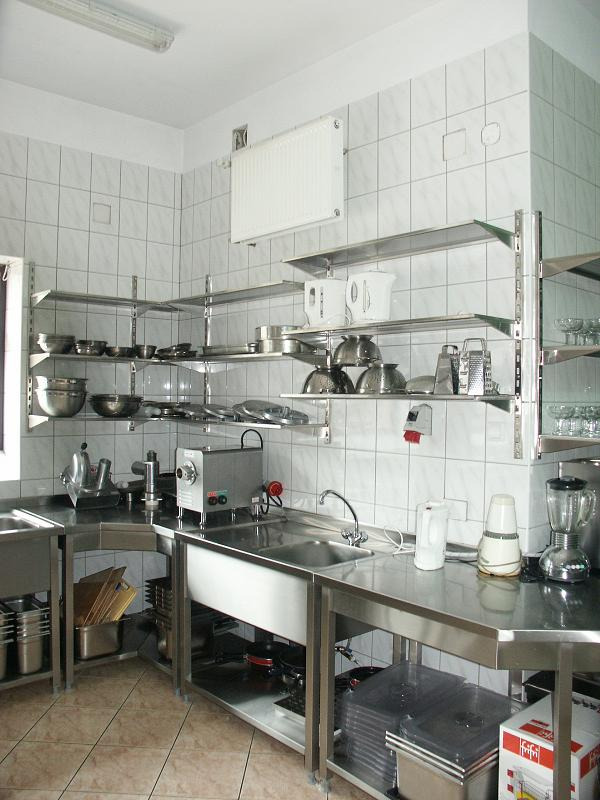 Wnętrze profesjonalnej kuchni z nierdzewnymi blatami, zlewozmywakami i półkami wypełnionymi naczyniami, widoczna maszynka do mielenia mięsa i inne urządzenia kuchenne.