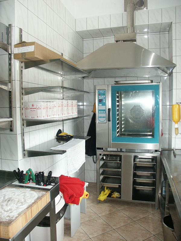 Profesjonalny piec konwekcyjny z okapem w kuchni restauracyjnej, widoczne półki z akcesoriami i deską do krojenia.