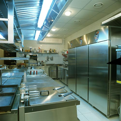 Profesjonalna kuchnia ze stali nierdzewnej z trzema dużymi lodówkami, widoczne blaty robocze, okap, oświetlenie i wyposażenie kuchenne.