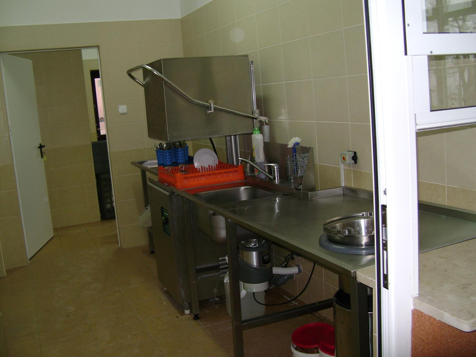 Profesjonalna zmywarka tunelowa ze stali nierdzewnej w lokalu gastronomicznym, z widocznym koszem z umytymi naczyniami, zlewem i pojemnikiem na odpady pod blatem.