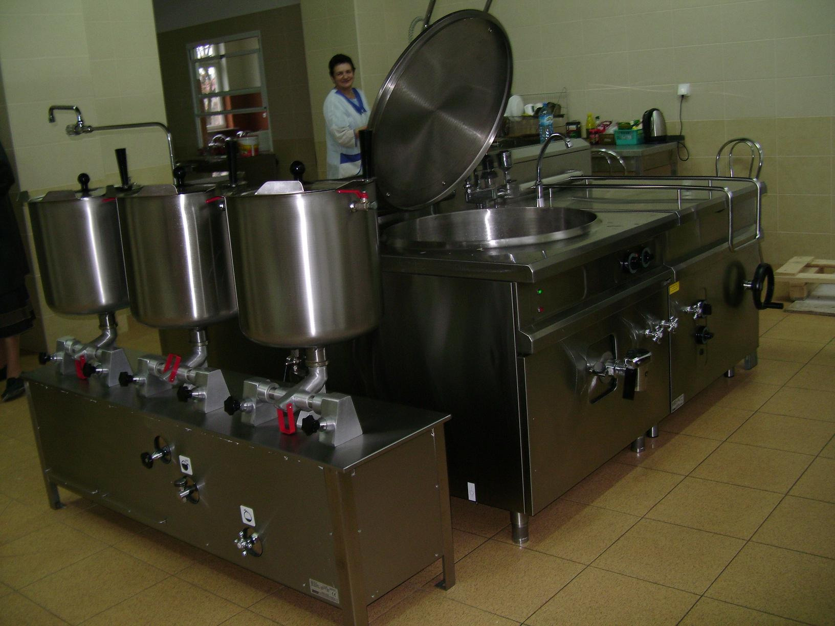 Profesjonalna kuchnia ze stali nierdzewnej: kotły warzelne i stół warzelny z otwartą pokrywą, widoczna osoba w fartuchu w tle.