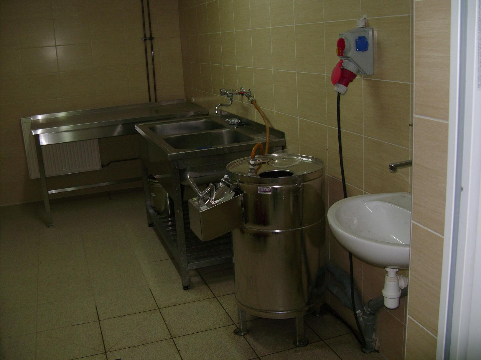 Wnętrze zaplecza kuchennego z metalowym stołem, dwukomorowym zlewem, okrągłą maszyną ze stali nierdzewnej z kranem i białą umywalką, przyłącze elektryczne na ścianie, wyłożone beżowymi płytkami.