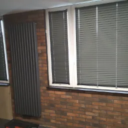 Szare, poziome osłony okienne harmonijkowe zamontowane w dwóch oknach z białymi ramami, na tle ściany z cegły i ciemnoszarego grzejnika.