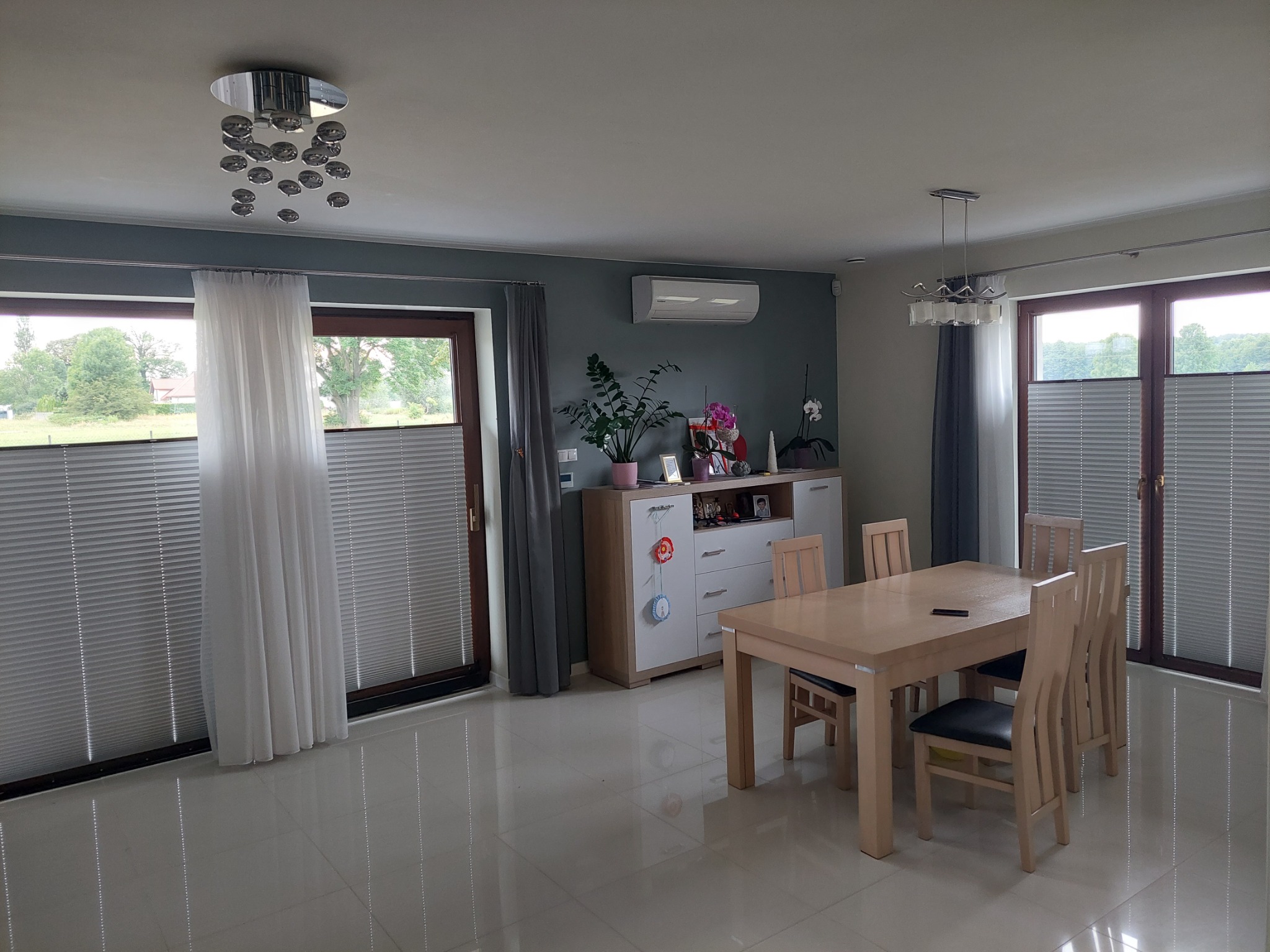 Jasne wnętrze jadalni z widocznymi oknami przesłoniętymi systemem zaciemniającym typu harmonijka w kolorze szarym, z białymi zasłonami po bokach okien, na podłodze błyszczące płytki.