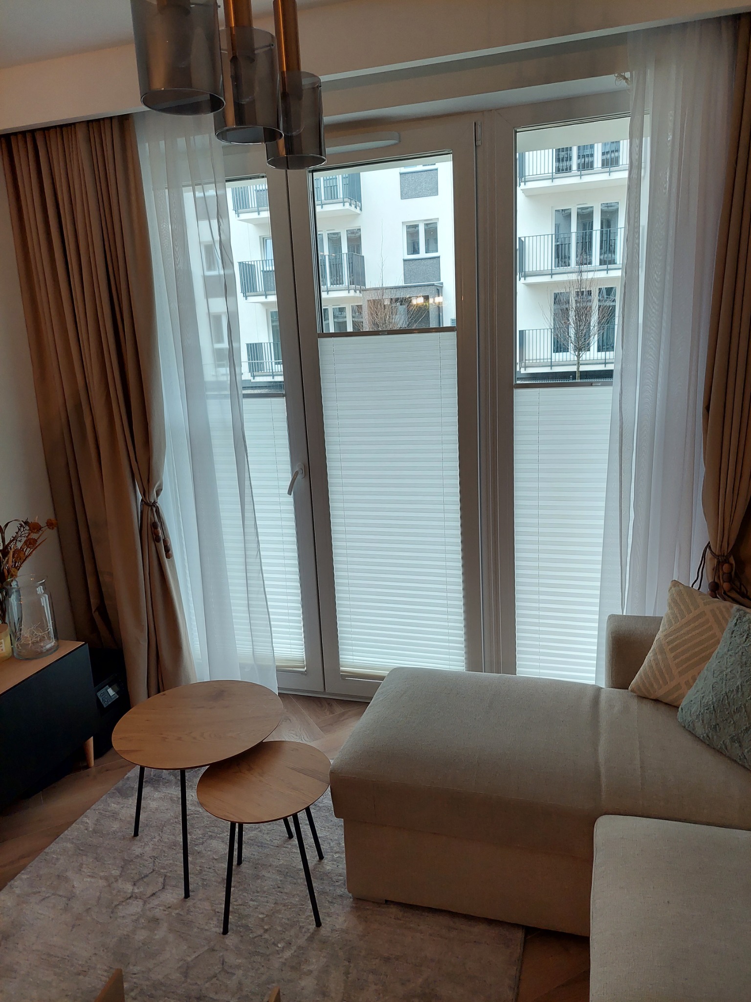 Jasne wnętrze salonu z oknami przesłoniętymi białymi, harmonijkowymi osłonami okiennymi, częściowo zasłoniętymi przez zwiewne, beżowe zasłony, z widokiem na budynek mieszkalny z balkonami.