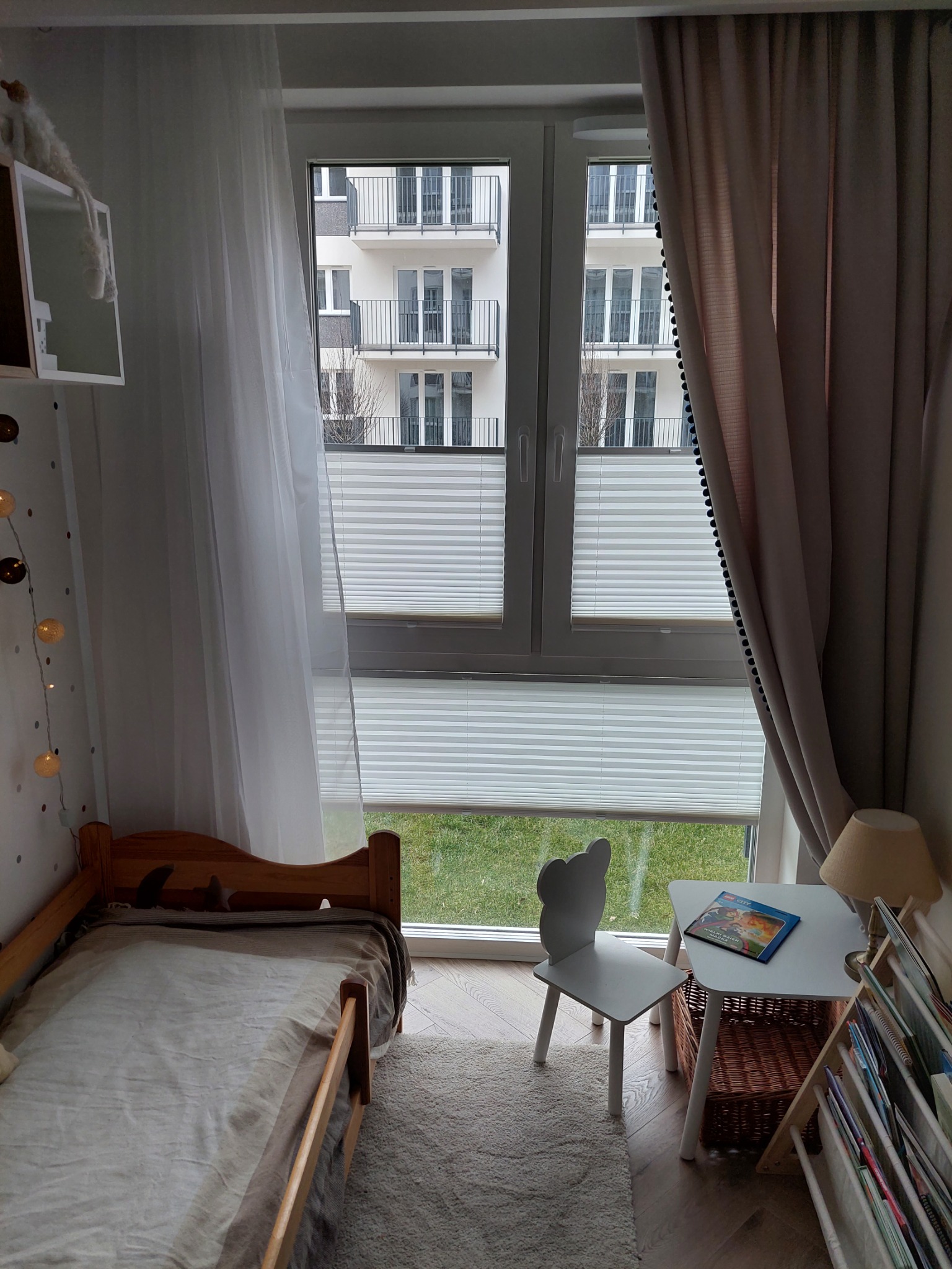 Dziecięcy pokój z oknem wyposażonym w białe, poziome, składane przesłony okienne, widok na budynek z balkonami