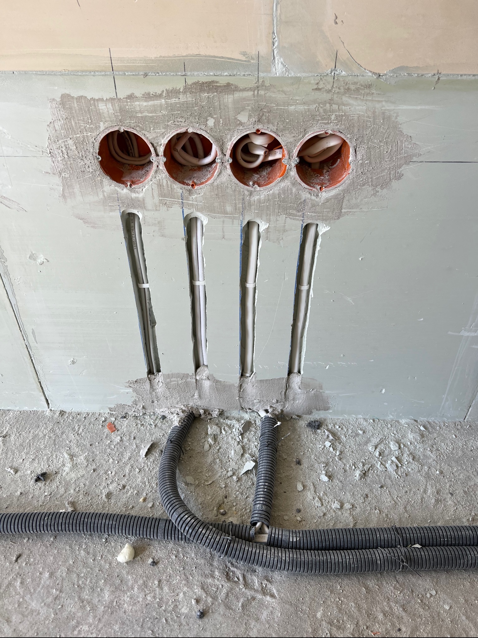 Ściana w trakcie prac wykończeniowych z widocznymi otworami na puszki elektryczne oraz pionowymi bruzdami z kablami w peszlach, na podłodze leżą dodatkowe peszle.