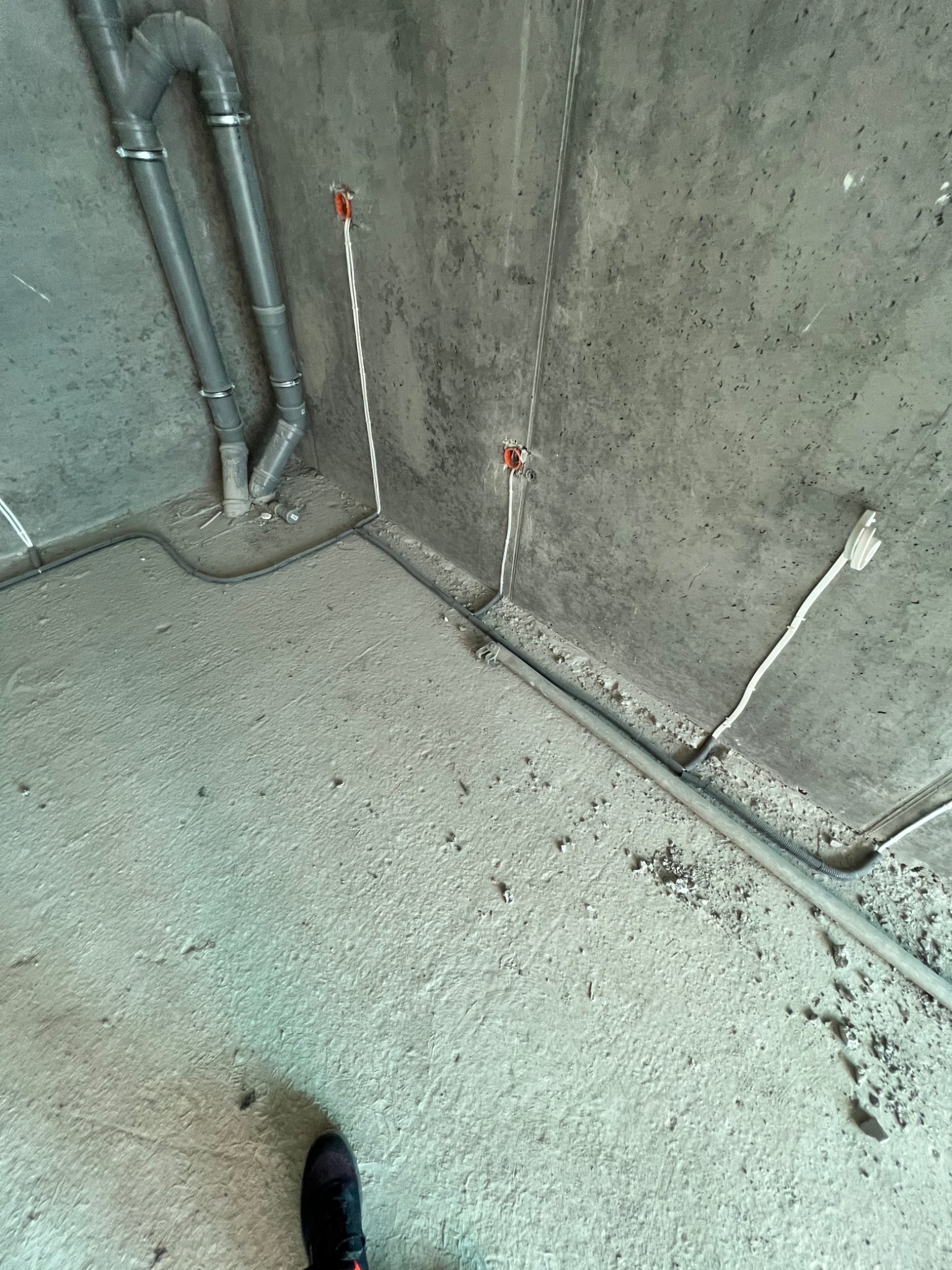 Surowy betonowy narożnik pomieszczenia w trakcie prac wykończeniowych z widocznymi rurami kanalizacyjnymi z metalowymi obejmami, przewodami elektrycznymi wystającymi ze ściany i kablami ułożonymi...