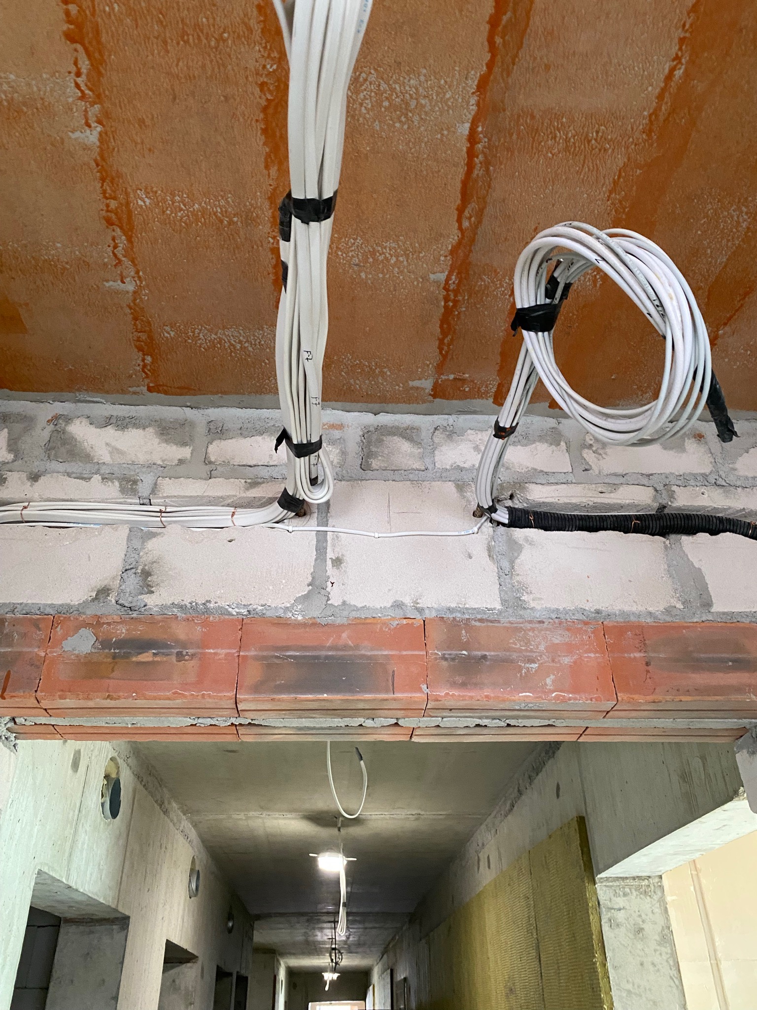 Wiązki białych przewodów elektrycznych przymocowane czarną taśmą do surowego sufitu z pustaków ceramicznych i ściany z białego betonu komórkowego w trakcie instalacji.