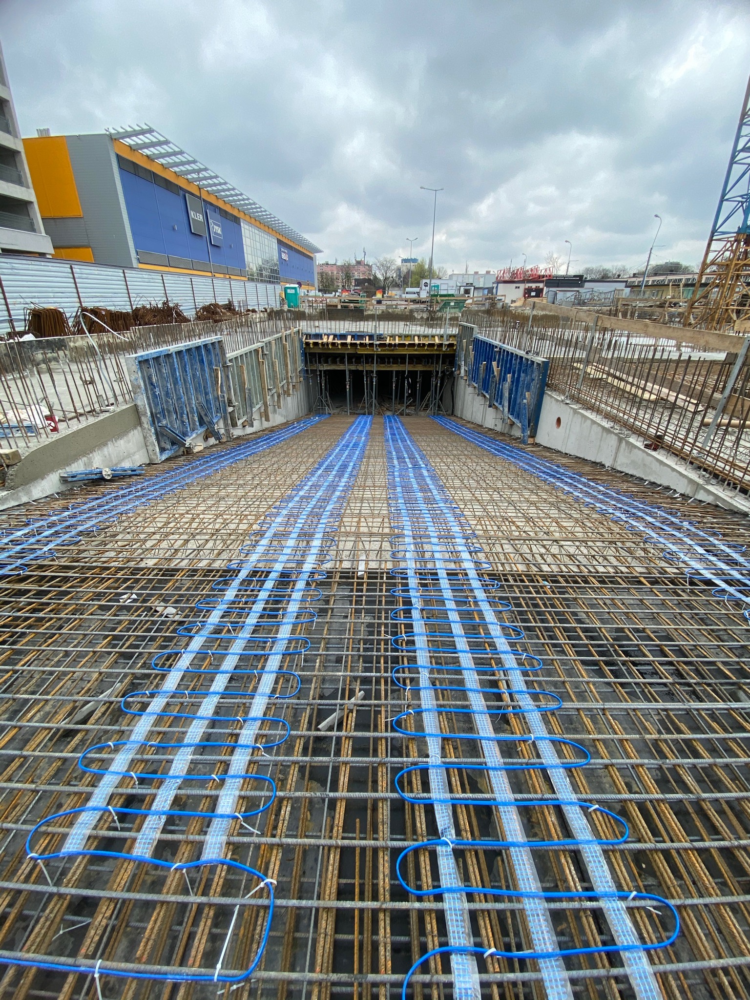 Widok z góry na konstrukcję zbrojeniową z przewodami grzewczymi ułożonymi w pętle na budowie tunelu drogowego, w tle widoczny budynek handlowy pod pochmurnym niebem.