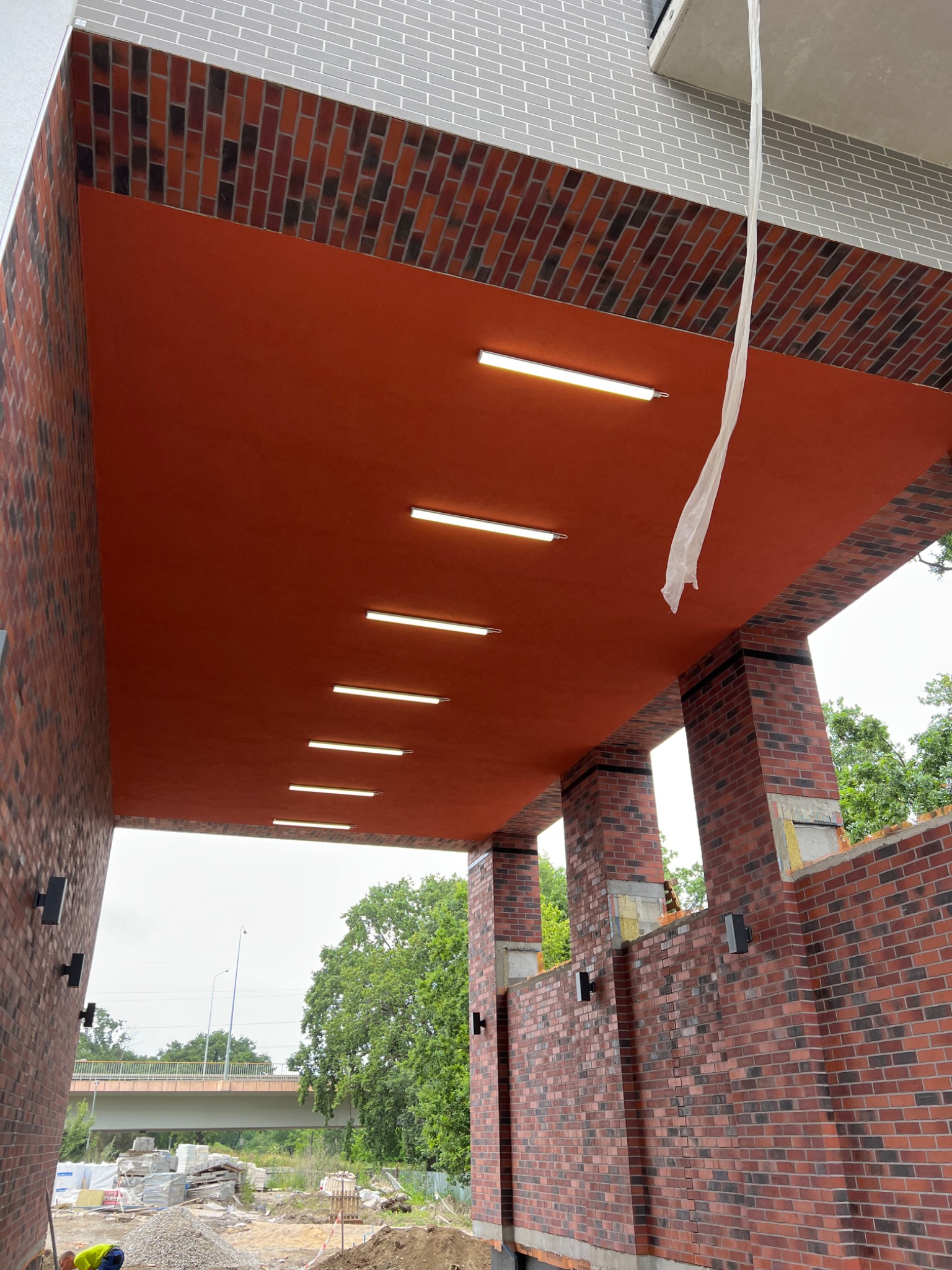 Zadaszenie z cegły klinkierowej z zamontowanym rzędem lamp liniowych LED, widok na most w tle, plac budowy z materiałami budowlanymi.