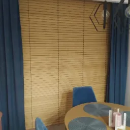 Drewniane żaluzje poziome w naturalnym odcieniu drewna, uzupełnione niebieskimi zasłonami i nowoczesną lampą wiszącą nad okrągłym stołem z niebieskimi podkładkami