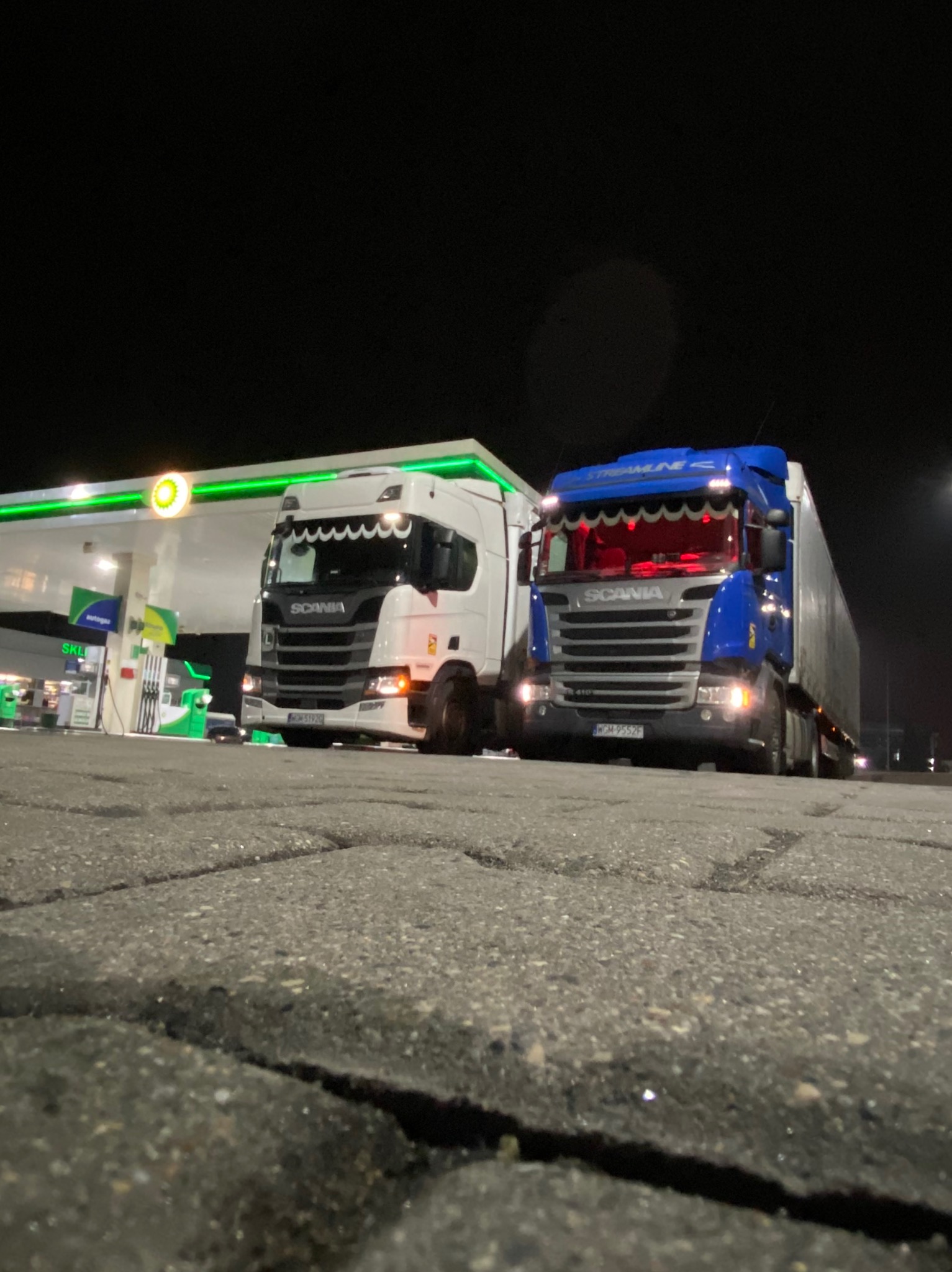 Dwa samochody ciężarowe marki Scania, biały i niebieski, stoją na stacji benzynowej nocą, oświetlone światłami stacji i reflektorami, widok z niskiej perspektywy.