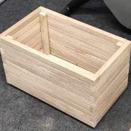 Drewniana skrzynka wykonana z listewek, widoczne łączenia na rogach i wewnętrzne wzmocnienie, surowe drewno, szary dywan w tle.