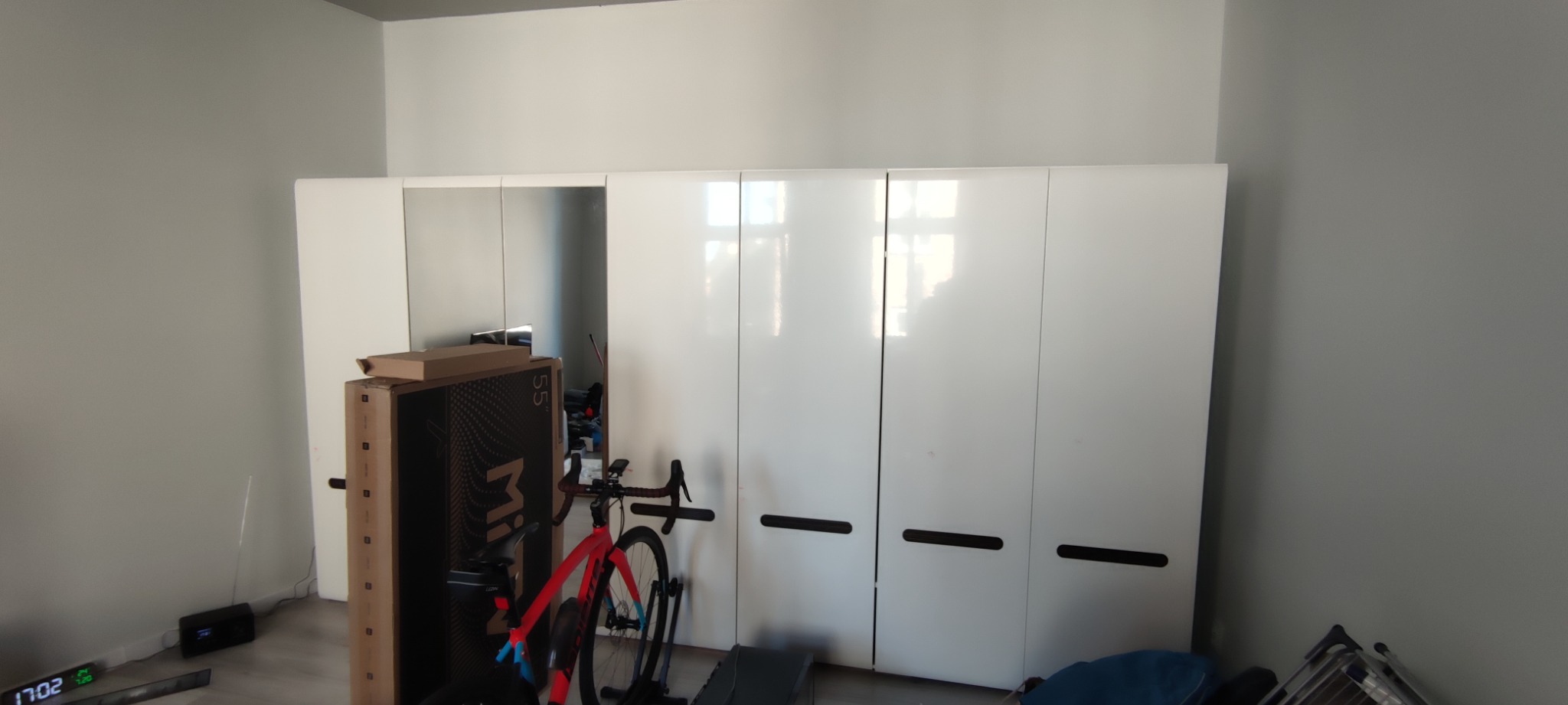 Biała szafa przesuwna z lustrzanymi drzwiami, częściowo zasłonięta kartonem po telewizorze i rowerem treningowym w pokoju o szarych ścianach.