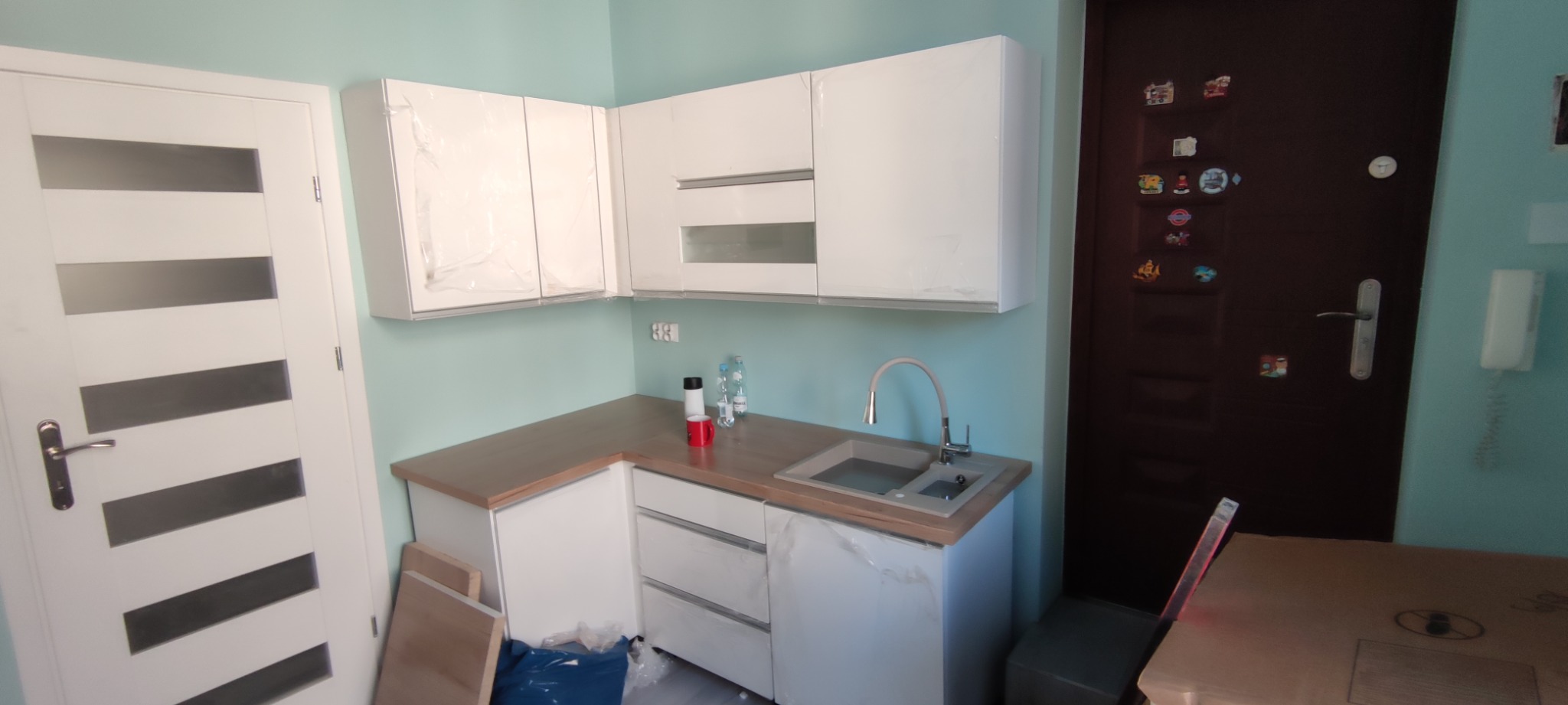 Nowa kuchnia z białymi szafkami, niektóre jeszcze w folii ochronnej, szary zlew i brązowy blat, białe drzwi z poziomymi przeszkleniami po lewej, brązowe drzwi wejściowe z magnesami po prawej...