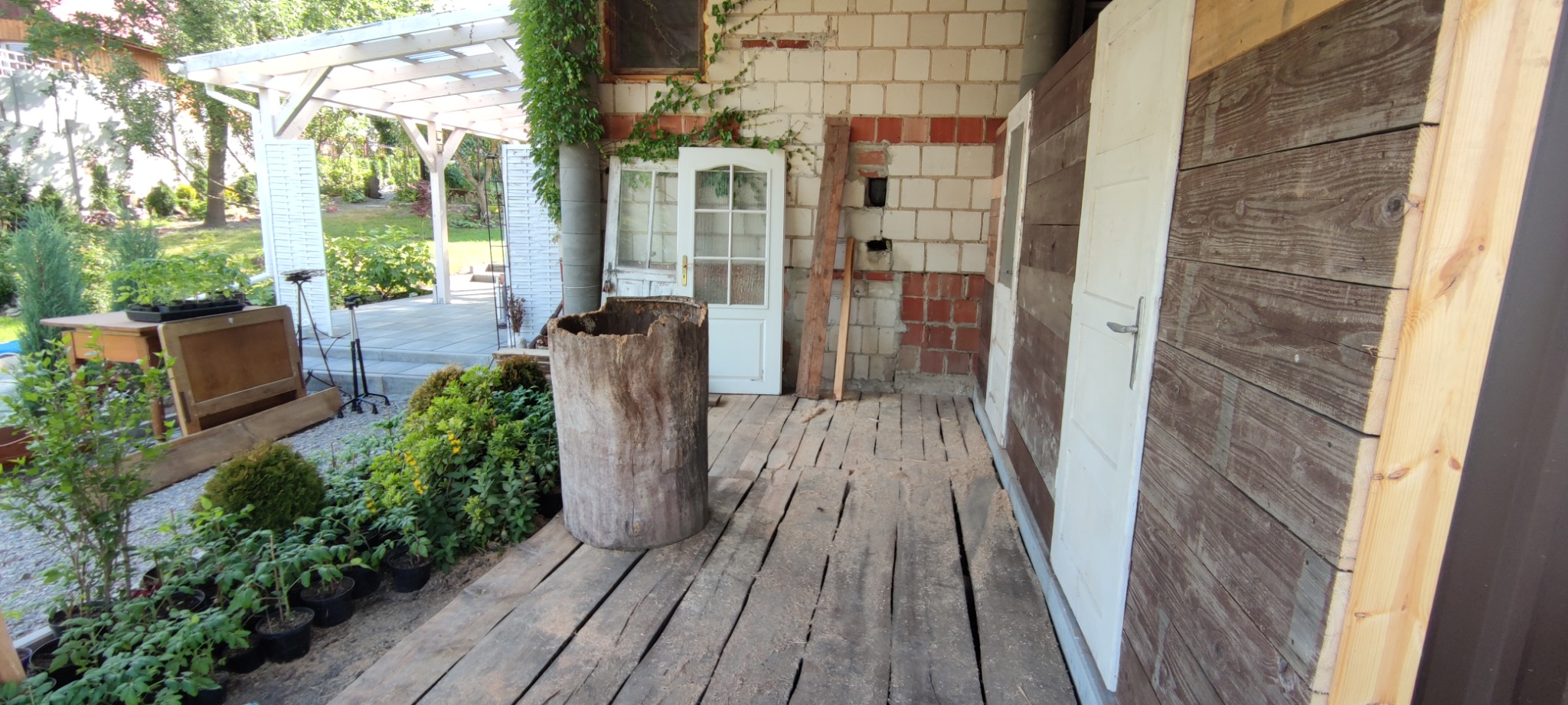 Taras z drewnianą podłogą, elementami starej stolarki okiennej i drzwiowej, oraz pniem drzewa pełniącym funkcję dekoracyjną.