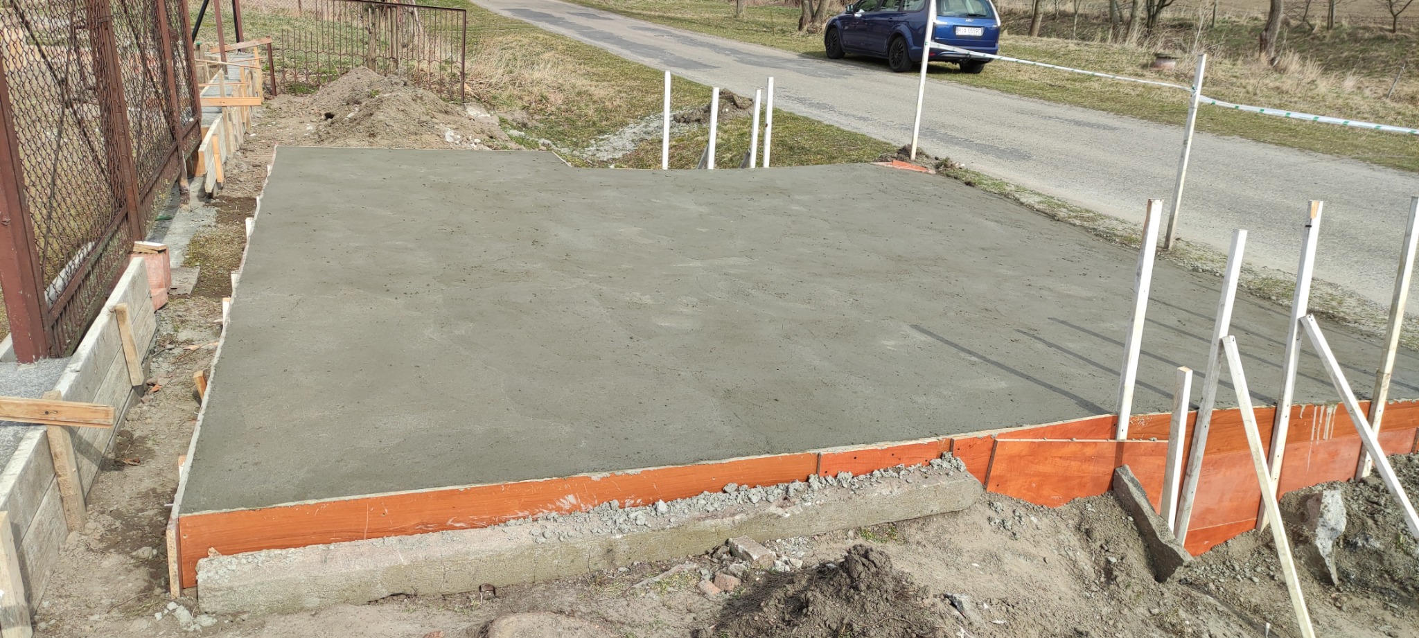 Świeżo wylana, gładka betonowa wylewka na przygotowanym podłożu z drewnianym szalunkiem, widoczne białe słupki i fragment drogi z samochodem w tle.