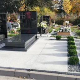 Dwa czarne, polerowane pomniki z wygrawerowanymi napisami na jasnoszarym granitowym podłożu, otoczone krzewami i kamieniami na cmentarzu w słoneczny dzień.